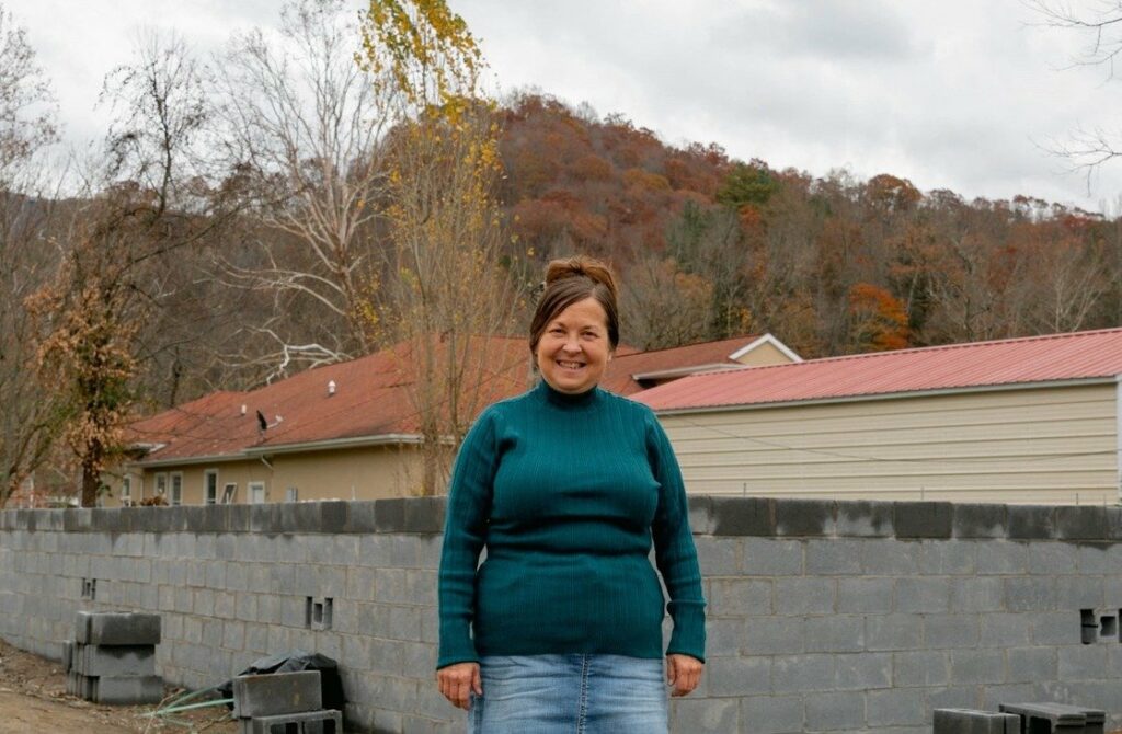 A woman stands in front of a partially constructed house, smiling and dressed in a teal sweater and denim skirt. She is surrounded by a concrete block wall, with building materials and the beginnings of a house structure in the background. The hillside behind her shows fall foliage, adding warmth to the scene. The image conveys hope and progress in rebuilding.