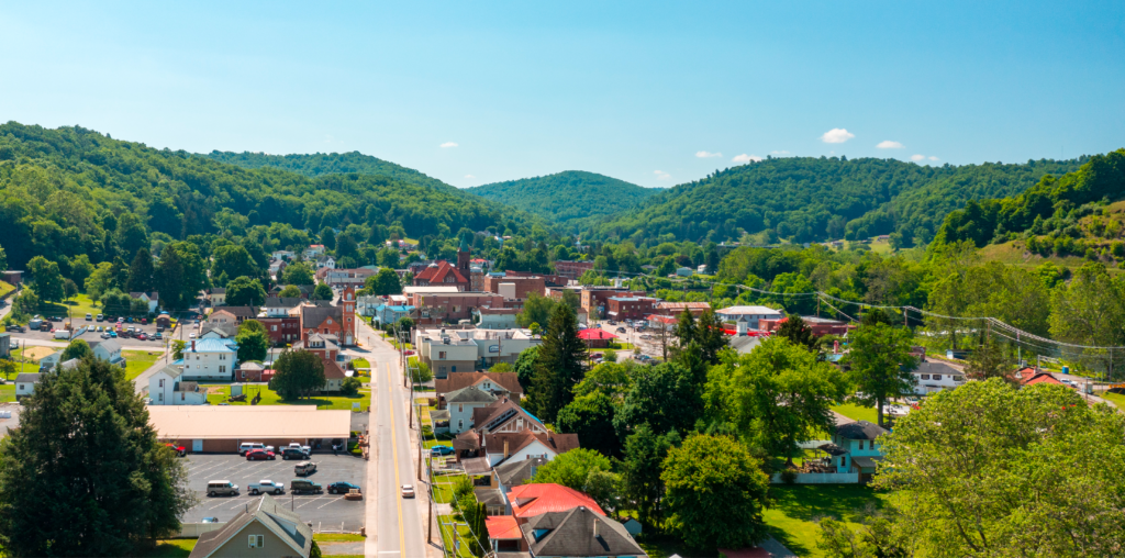 A small Appalachian town sits nestled in a lush green valley, with tree-covered hills rising in the background under a clear blue sky. The town features a mix of homes, historic buildings, and a central road lined with cars and community activity.