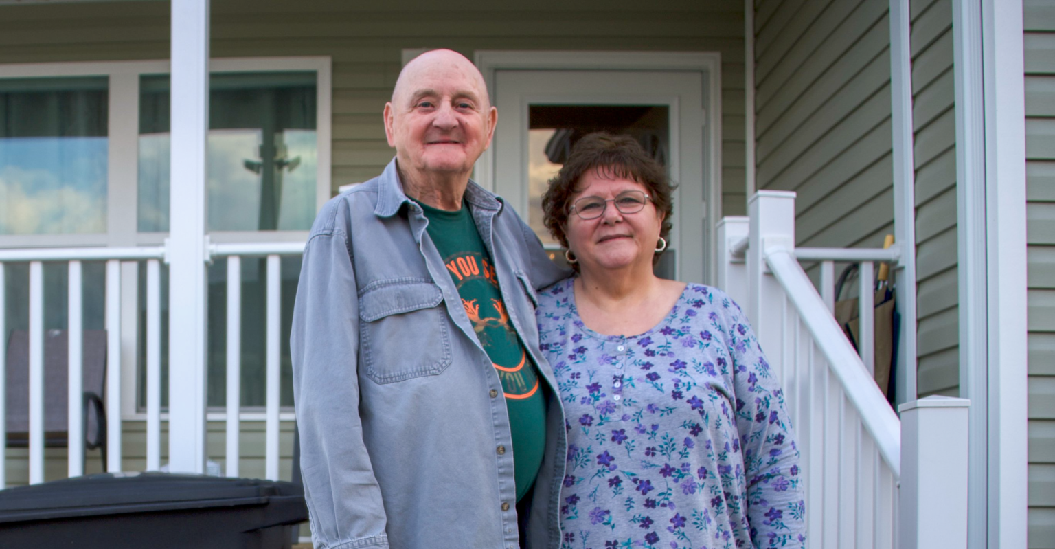 This image shows an older couple standing proudly in front of their home, smiling and arm-in-arm. They’re posed on the front porch, framed by white railings and a clean, well-maintained house exterior. Their expressions radiate warmth, stability, and a sense of belonging—capturing a moment of pride and comfort in their place of residence.