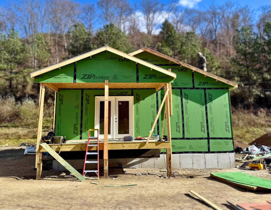 A small house under construction features green ZIP System sheathing on the exterior walls and exposed framing for the front porch. A red-and-white ladder leans against the unfinished deck, and construction tools and materials are scattered around the dirt worksite. A worker is visible on the roof, and leafless trees and pines stand in the background under a blue sky.