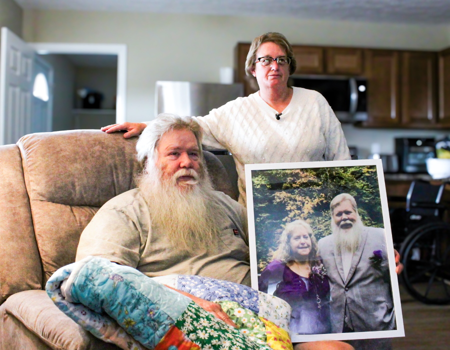 A man with a long white beard sits in a recliner covered by a colorful patchwork quilt, while a woman stands beside him with her hand resting on his shoulder. The man holds a large framed photo of the couple taken in an outdoor setting, both smiling and dressed formally. Behind them is a kitchen with wooden cabinets and stainless steel appliances, and a wheelchair is partially visible in the background, suggesting the couple's resilience and support for one another in their home.