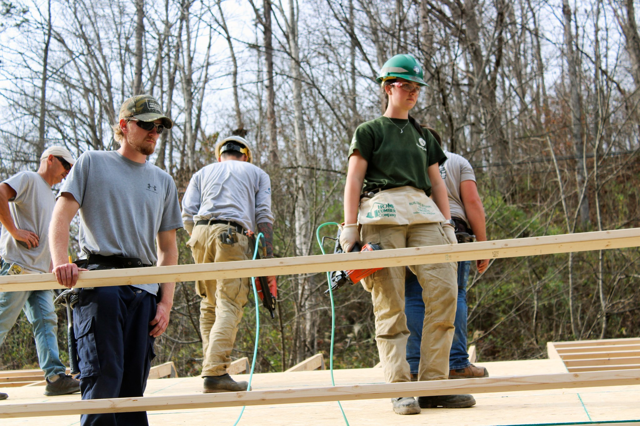 A group of construction workers and volunteers work together on a building site, carrying wooden beams and using tools. Surrounded by leafless trees, they focus on the task at hand, showcasing teamwork and progress on a new home or structure.