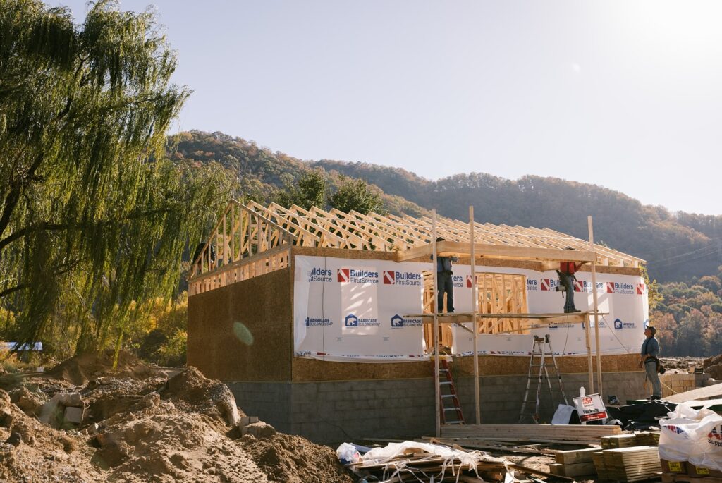 Construction workers build the frame of a new house, surrounded by lumber, tools, and scaffolding on a clear, sunny day. The structure is set against a backdrop of tree-covered hills, signaling progress and hope in a rural or mountainous community.