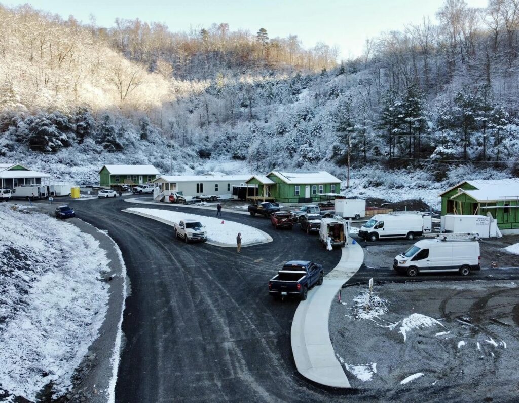 A construction site in a snowy mountain valley shows multiple homes in progress, with green sheathing visible on several structures. Work trucks and construction crews are active on the freshly paved road, surrounded by frosted trees and hills.