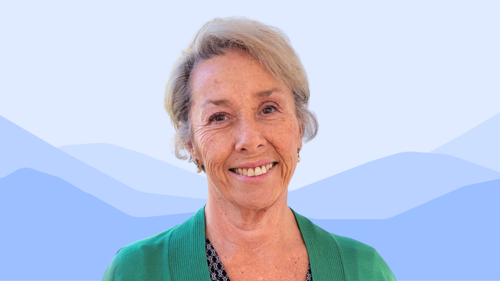 Lindy Turner, a smiling woman with short, light gray hair, wears a green cardigan over a patterned top, standing in front of a soft blue background with abstract mountain shapes.