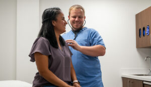 A healthcare provider in blue scrubs smiles while using a stethoscope to listen to a patient's heartbeat. The patient, seated and also smiling, appears relaxed in a clean and modern medical exam room.