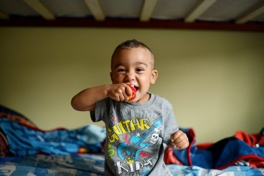 A joyful toddler sits on a colorful bed, smiling and playfully biting a red toy while wearing a gray "Guitar Kids" t-shirt. The room has a cozy, lived-in feel with blankets and bedding scattered behind him.