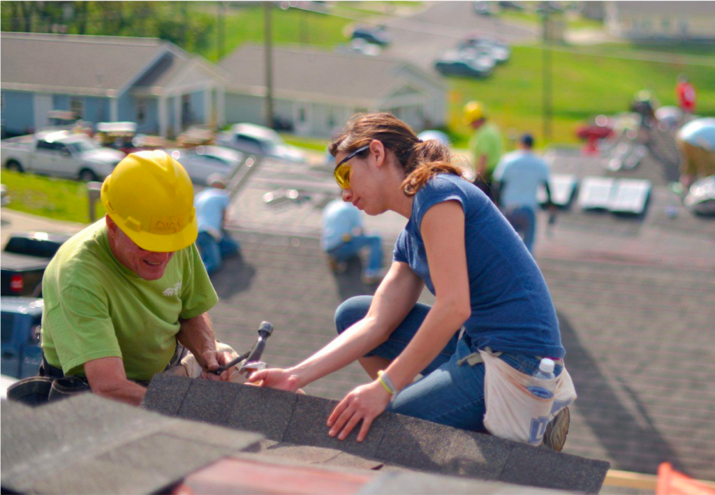 A man in a yellow hard hat and a woman wearing safety glasses work together on a rooftop, installing shingles as part of a group construction effort. Other volunteers can be seen working on nearby rooftops in the background.