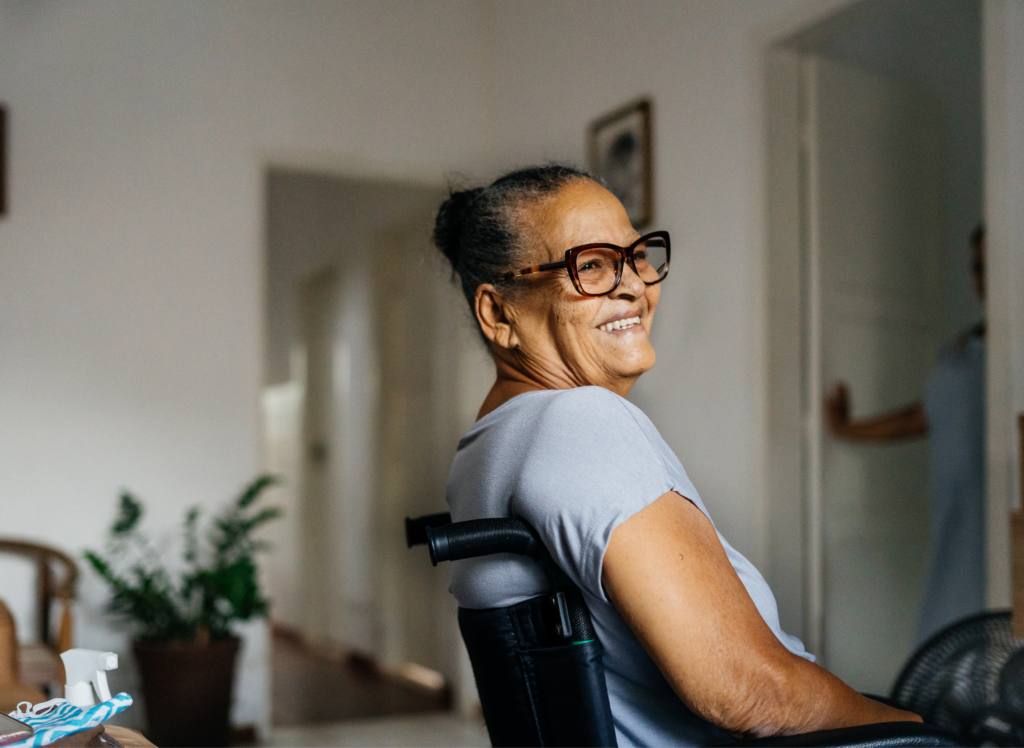 An older woman with glasses smiles while sitting in a wheelchair inside a warmly lit home. She appears relaxed and content, with a houseplant and a hallway visible in the background.