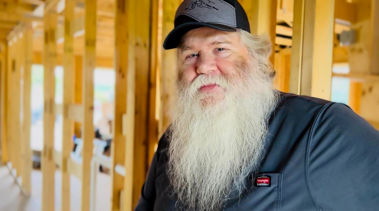 A man with a long white beard and a black cap smiles softly while standing inside a house under construction. Wooden framing surrounds him, and natural light filters through the unfinished walls.