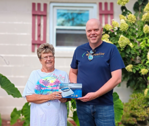 A smiling older woman and a man stand outside a house holding a weather alert radio box. The woman wears a beach-themed shirt, and the man wears a "Creative Passion, Inc." T-shirt.