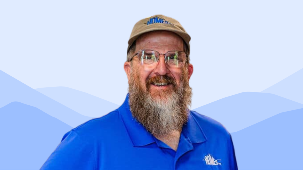 Seth Long smiles while wearing a tan cap, glasses, and a blue polo shirt with the HOMES Inc. logo, standing in front of a light blue background with stylized mountain shapes.