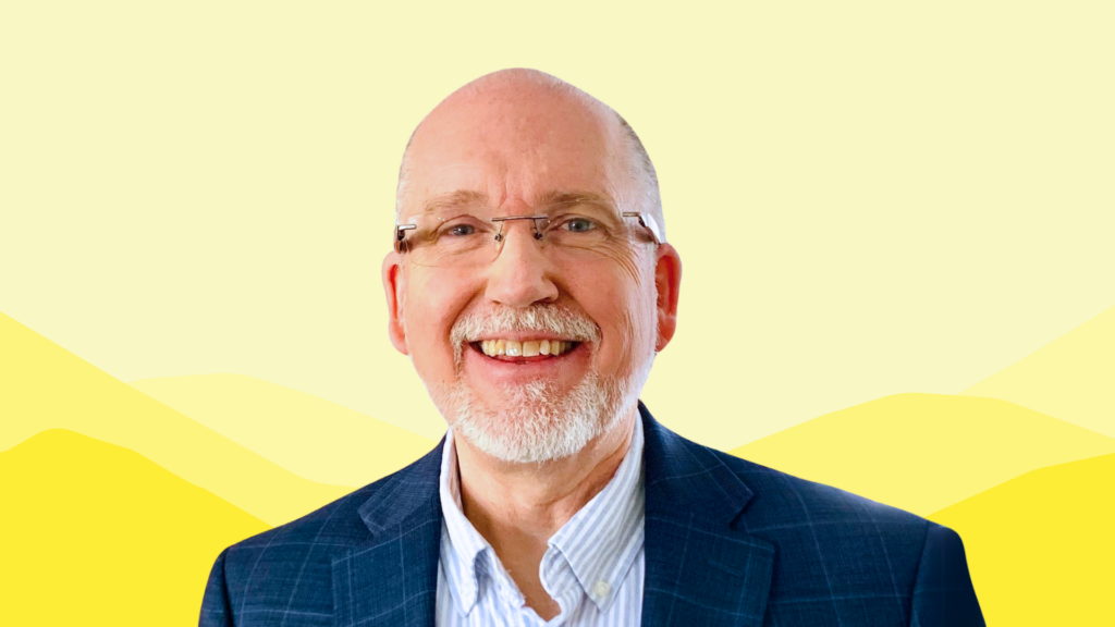 Chris Mullett, a smiling man with glasses, a white beard, and a plaid navy blazer, stands in front of a pale yellow background featuring stylized mountain shapes.