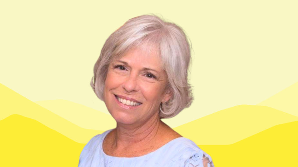 Mary Ellen Judah, a smiling woman with short, light gray hair, wears a pale blue top, standing in front of a yellow background with soft, stylized mountain shapes.