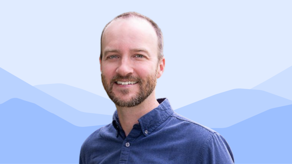 A smiling man with a beard and short hair wears a blue button-down shirt, standing in front of a light blue background with stylized mountain shapes.