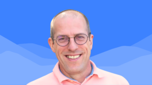 A smiling man wearing glasses and a light pink collared shirt stands in front of a blue background with abstract mountain shapes.
