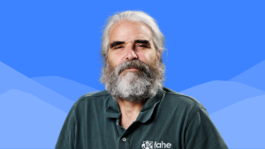 Scott McReynolds stands against a blue background with stylized mountain shapes, wearing a dark green Fahe polo shirt and sporting long gray hair and a full beard.
