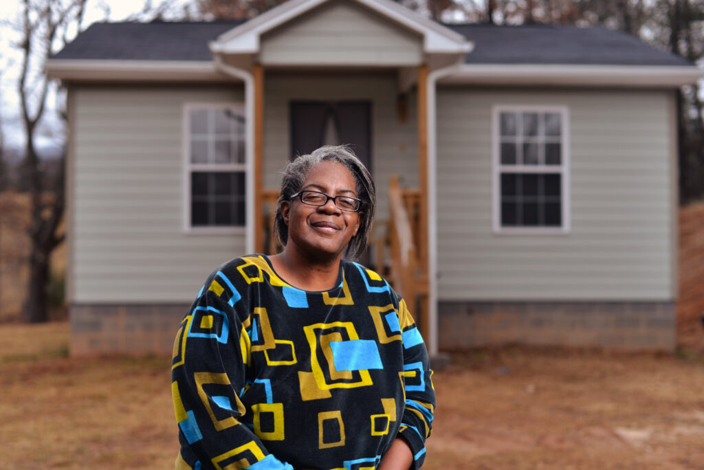 A woman wearing a colorful geometric-patterned sweater smiles confidently while standing in front of a newly built house with light gray siding and white trim. The setting suggests a sense of pride and accomplishment in a peaceful, rural neighborhood.