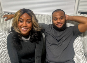 A smiling couple sits closely together on a cozy, textured couch with patterned pillows. Their relaxed posture and joyful expressions reflect warmth, comfort, and a sense of home.