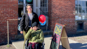 A smiling woman stands beside a young person in a wheelchair outside a brick building, near a colorful chalkboard sign that reads "Cafe OPEN NOW." Red and blue balloons add a festive touch, suggesting a welcoming and inclusive community space.