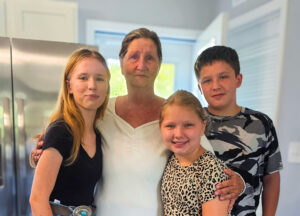 A woman stands in a brightly lit kitchen with three children, all smiling warmly for the photo. Their close embrace and relaxed expressions reflect a sense of family, love, and togetherness in their home.