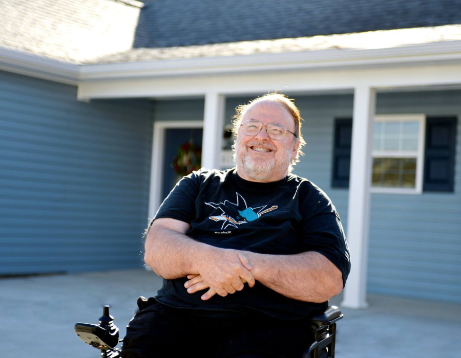 A man smiling outside of a blue house on a sunny day.