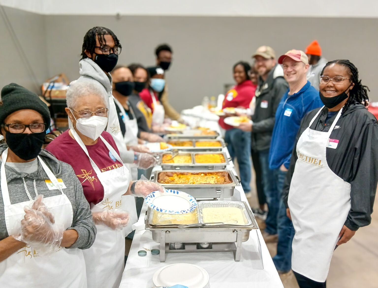 A group of volunteers, wearing aprons and masks, stand behind a serving table filled with trays of food. They are smiling as they prepare to serve meals, with a variety of dishes visible, such as casseroles and baked goods. The volunteers range in age and ethnicity, and they are all wearing gloves as they help with the meal service. The background shows additional people in line, and the setting appears to be a community meal or event at a large indoor space.