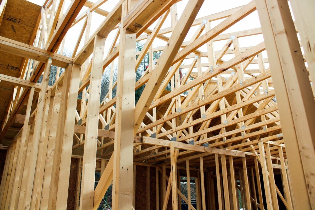 A close-up view of the wooden framework of a house under construction, showing a grid of beams and trusses. The image captures the intricate structure of the walls and ceiling, with metal connectors visible at the joints. The natural light is coming through the top, illuminating the raw wood.