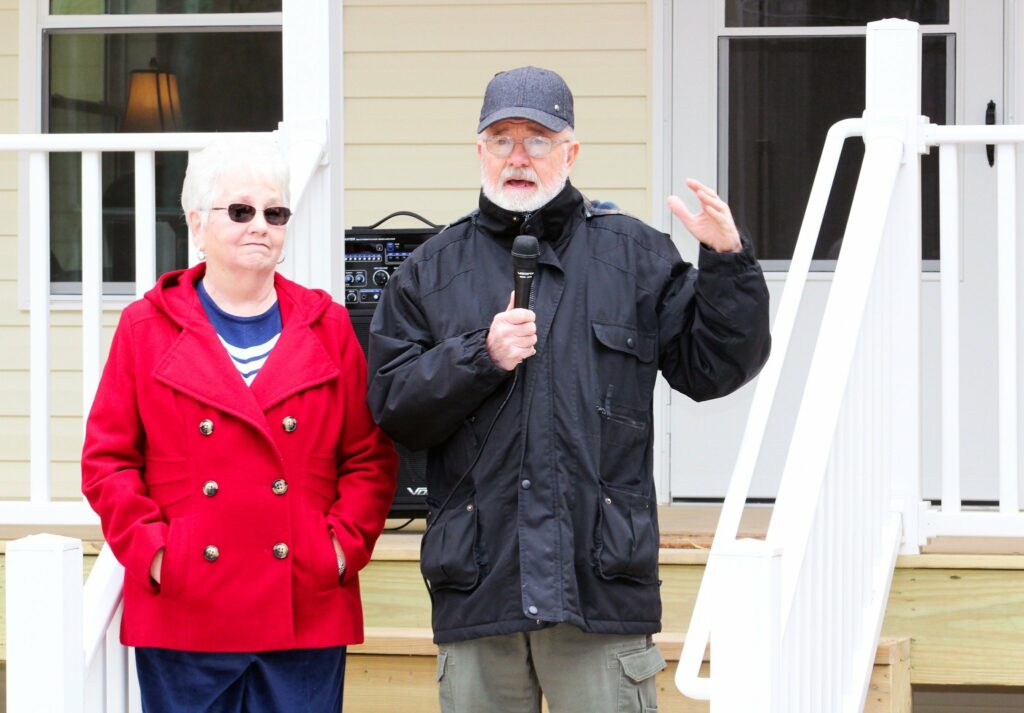 A man is standing on the porch of a newly built house, holding a microphone and speaking. He has a white beard and is wearing a black jacket and a dark cap. Next to him stands a woman wearing sunglasses and a red coat with a striped shirt underneath, looking slightly to the side. Behind them, a speaker can be seen, and part of the house's porch and railing are visible.