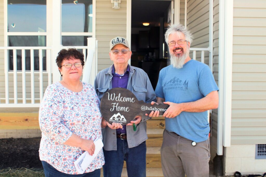 A woman and a man, both smiling, stand outside a newly built home holding a large "Welcome Home" sign. The man is wearing a gray cap and a blue shirt, and the woman is wearing a floral blouse. They are joined by another man, also smiling, who is wearing a blue t-shirt and holding the sign. The house's front porch is visible in the background, with a few steps leading up to the door. The sign features the logo for Housing Development Alliance (@hdahome).