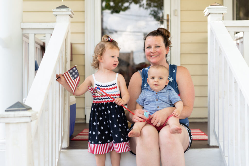 A smiling woman sits on a front porch holding a baby, while a young girl stands beside her waving an American flag. All three are dressed in patriotic red, white, and blue outfits, celebrating a summertime holiday.