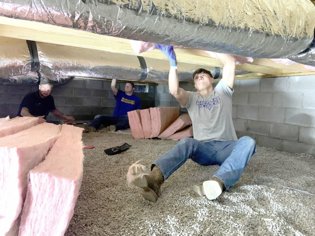 Three volunteers are working together under a house in the crawl space. One person is seated on the gravel floor while another is kneeling and installing pink insulation in the ceiling. A third volunteer is standing, holding up ductwork. All volunteers are wearing gloves and work clothing. The scene is dimly lit, and rolls of insulation are scattered on the ground.
