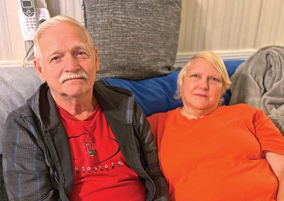 A middle-aged couple sitting together on a couch. The man, wearing a red t-shirt with a logo and a black jacket, has a white mustache and short gray hair. The woman, wearing an orange t-shirt, has short blonde hair and a gentle expression. They are sitting closely together, looking directly at the camera.