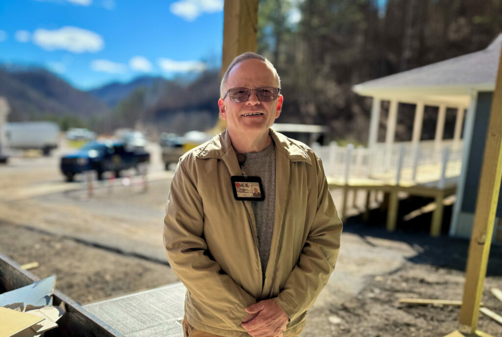 A man in a tan jacket and glasses stands smiling in front of a construction site on a sunny day, with mountains and newly built homes in the background.