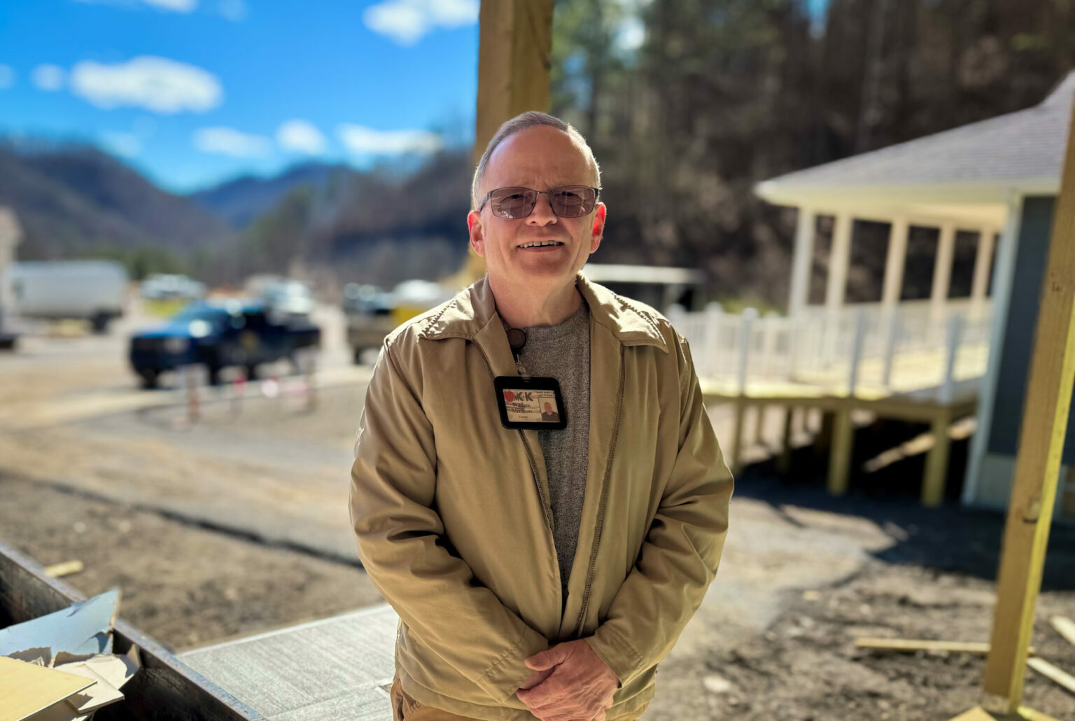 A man in a tan jacket and glasses stands smiling in front of a construction site on a sunny day, with mountains and newly built homes in the background.