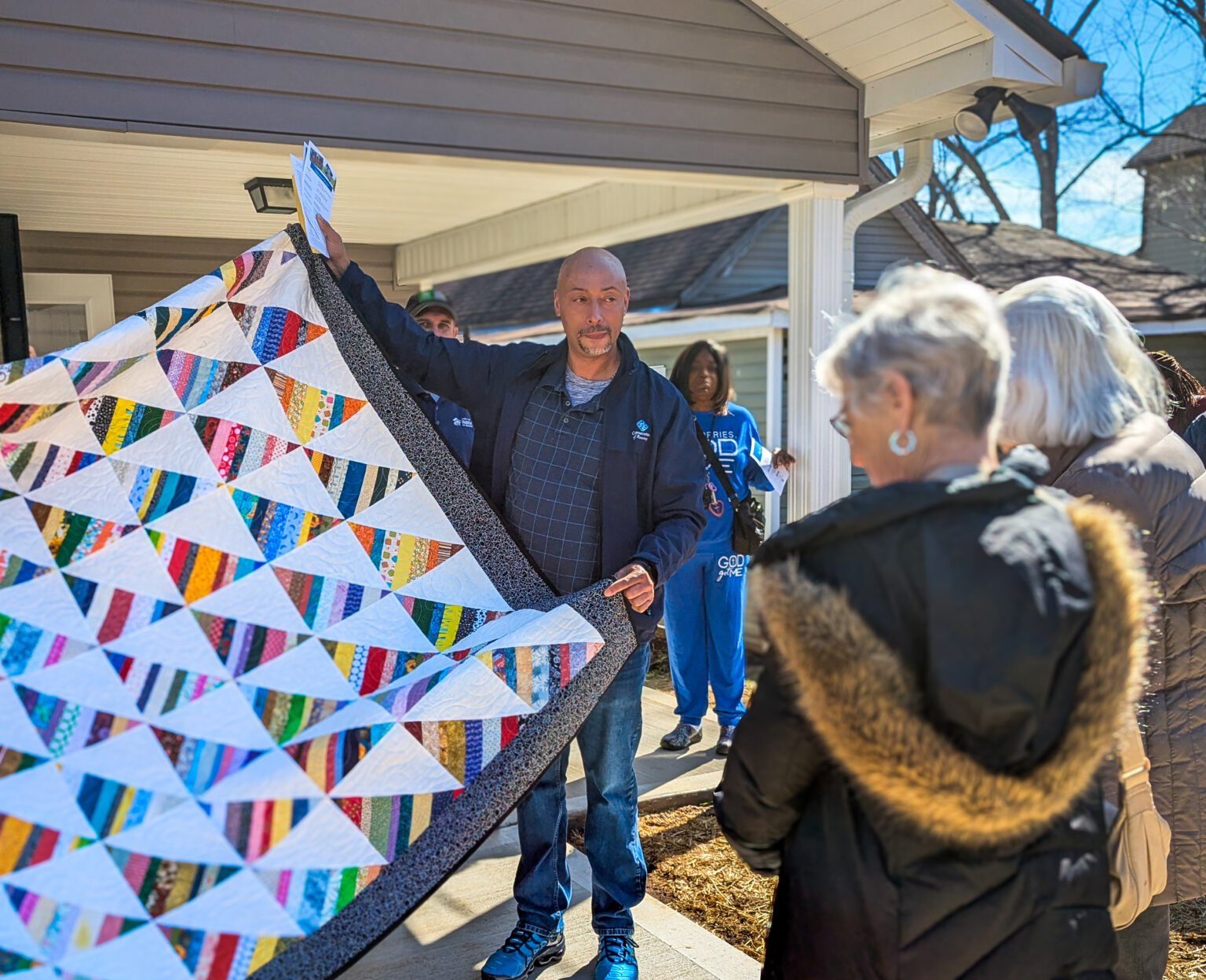A man stands in front of a house holding up a colorful patchwork quilt, addressing a small group of people gathered outside.