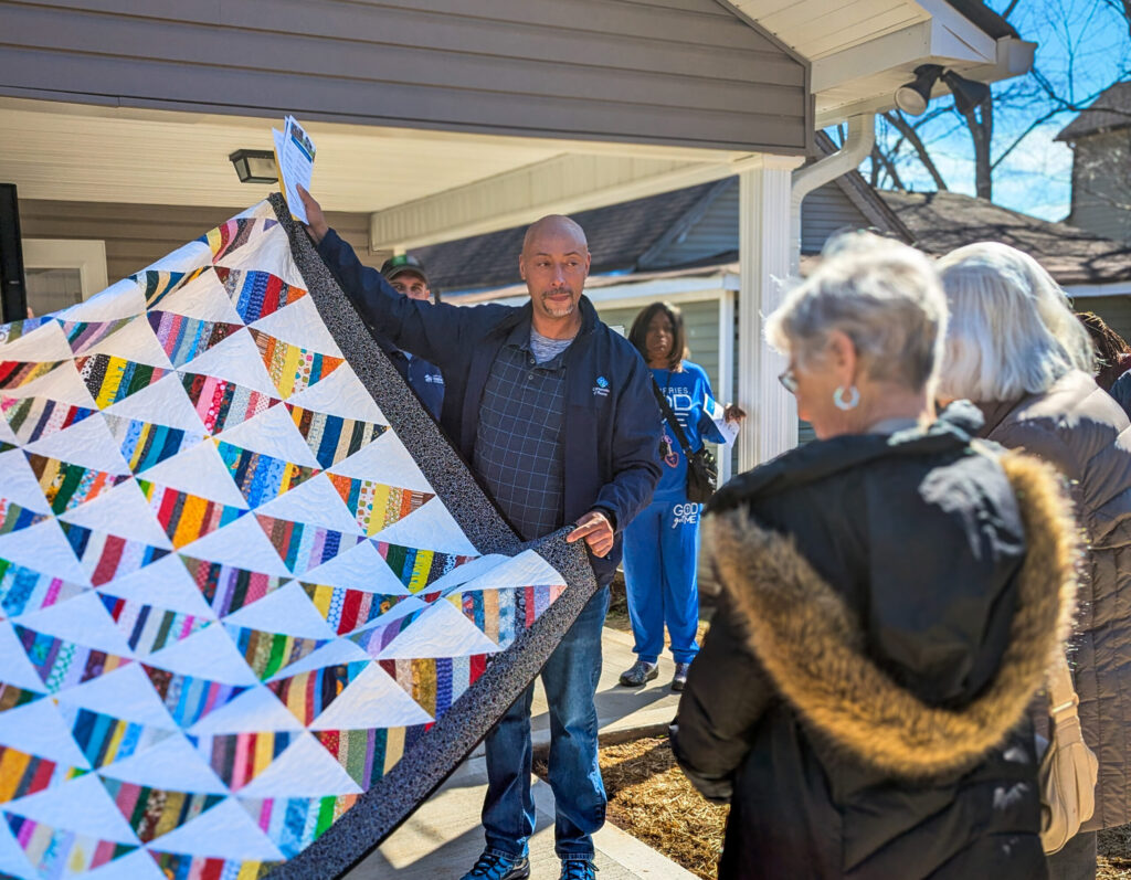 A man stands in front of a house holding up a colorful patchwork quilt, addressing a small group of people gathered outside.