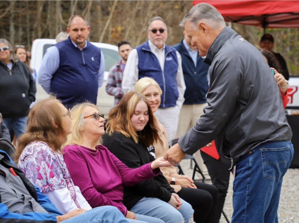 A man in a black jacket shakes hands with a woman seated in front of a small crowd. The woman, wearing glasses and a purple shirt, looks up at the man as she extends her hand. Behind them, a group of people stands, with several others sitting in chairs, watching the interaction. The setting appears to be an outdoor event, with trees and vehicles in the background. The image captures a moment of connection and community.