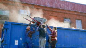 Three people, wearing work gloves and casual clothing, are lifting a wheelbarrow full of debris into a large blue dumpster. Dust rises from the contents of the wheelbarrow as they work together to unload it. The backdrop shows a red brick building with boarded-up windows.