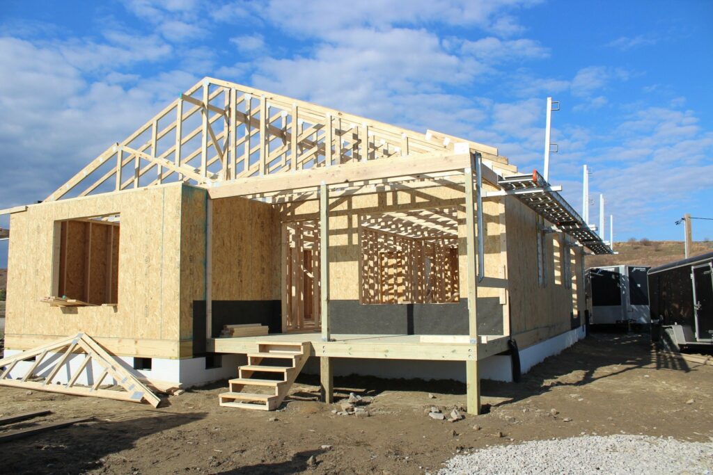 A partially constructed house with a wooden frame is shown against a blue sky with clouds. The house has a set of wooden stairs leading up to the entrance, and the roof trusses are being built. The walls are made of plywood, with an open window frame visible. In the background, a few trailers and piles of construction materials are scattered on the ground.