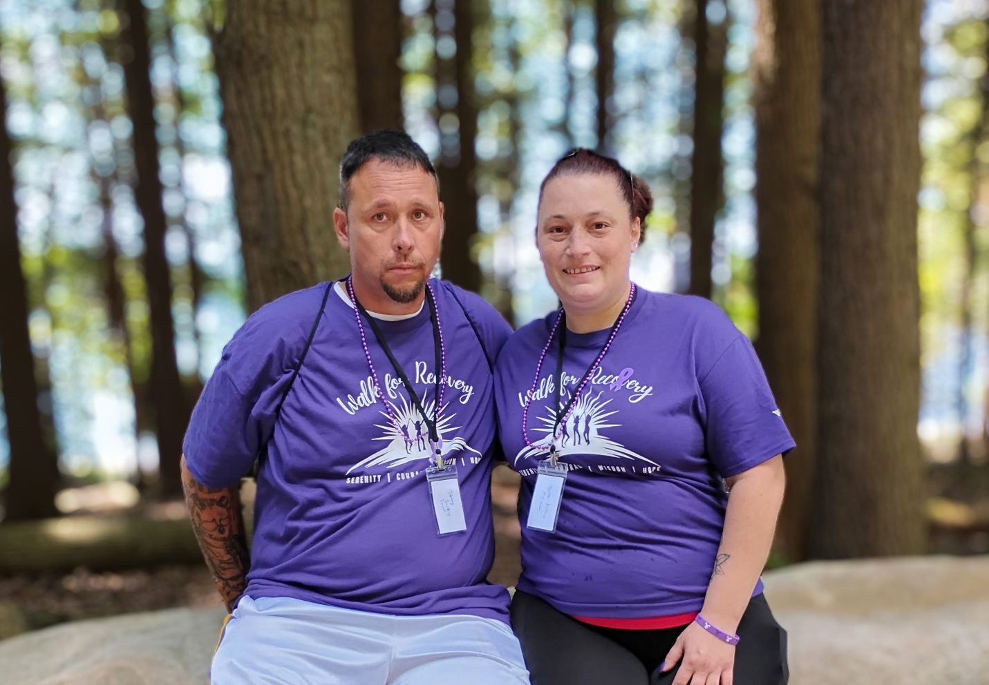 A man and woman sit close together on a large rock in a wooded area, both wearing matching purple “Walk for Recovery” t-shirts and participant badges.